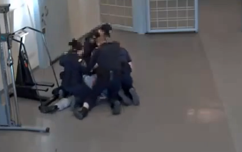 Photo of an inmate lying prone on the floor with four officers on top of him